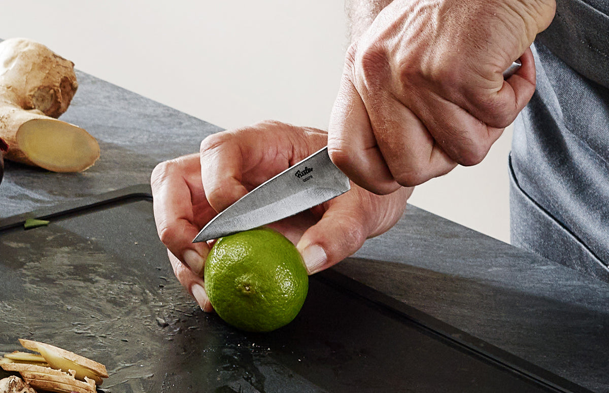 A person cuts a lime with a Fissler Essential® paring knife on a smooth stone surface; a fresh ginger root lies in the background.