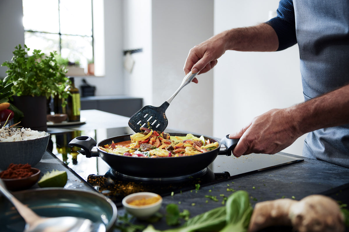 Cooking vibrant vegetables in a black Fissler Essential® Frying Pan with silicone spatula, fresh herbs on the counter.