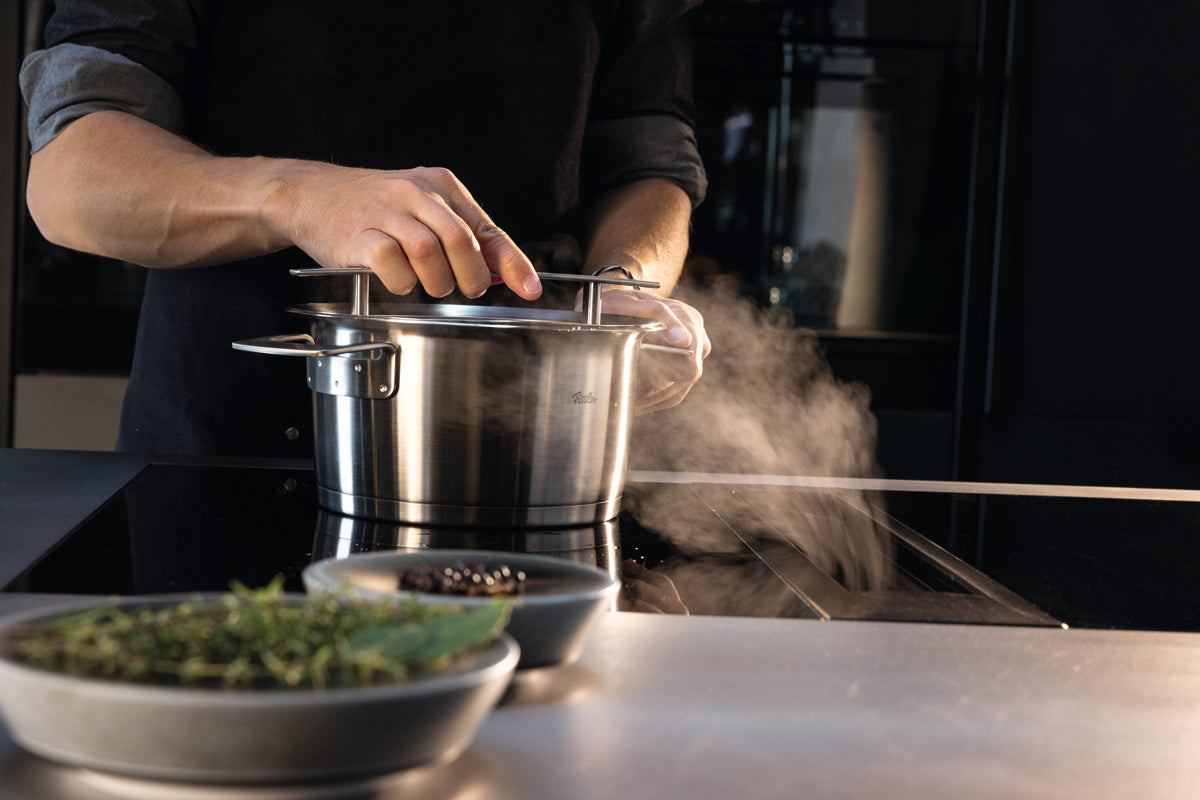 Discover Fissler precision: lifting the Phi Collection® stock pot lid, steam rises; fresh herbs and peppercorns nearby.