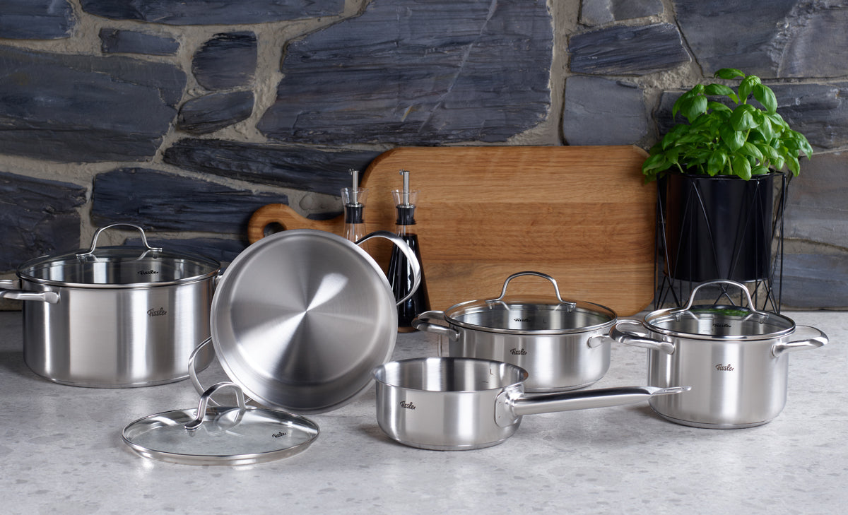 A set of Fissler San Francisco stainless steel pots and pans with lids, featuring precision-engineered German design, arranged on a kitchen counter in front of a stone wall.