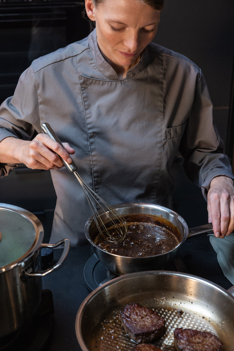 A chef in a grey uniform uses precision to whisk a sauce in a Fissler Original-Profi Collection® Sauteuse 20cm, reflecting our heritage of German-engineered quality, on a stovetop next to a pan containing seared steaks.