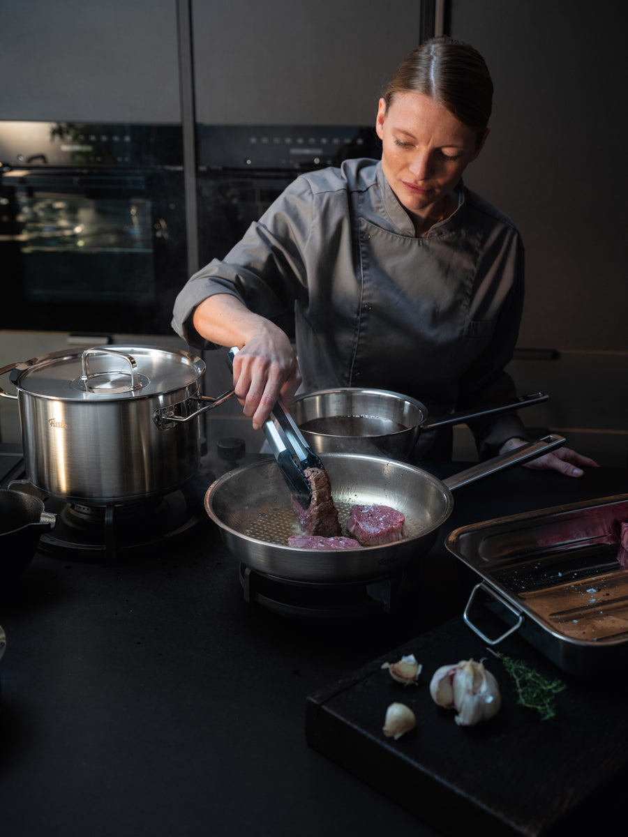 Ein Koch brät ein Steak in einer Pfanne der Fissler Original-Profi Collection®, umgeben von unserem hochwertigen Kochgeschirr, Kräutern und Knoblauch.