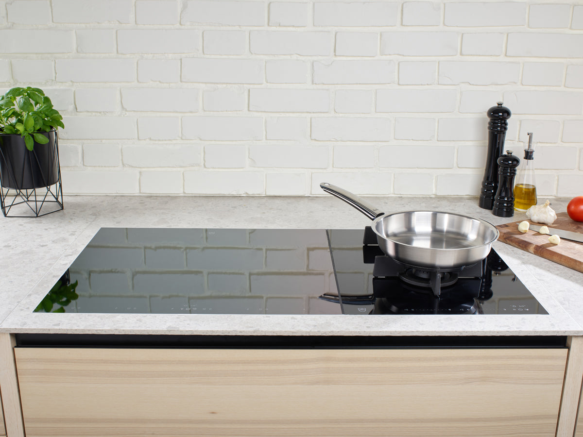 In einer modernen Küche kommt ein elegantes Induktionskochfeld aus schwarzem Glas mit dem 2-teiligen Pfannenset von Fissler zur Geltung: Ceratal® Classic-Pfanne 28 cm und Pure-Pfanne 24 cm. Das präzise gefertigte, langlebige Kochgeschirr von Fissler – seit 1845 in Deutschland hergestellt – steht neben frischen Kräutern in einem Pflanzkübel, einer Pfeffermühle, einer Olivenölflasche, Knoblauchzehen, rustikalem Brot und einer reifen Tomate auf einem Holzschneidebrett und steht für zeitloses Design und