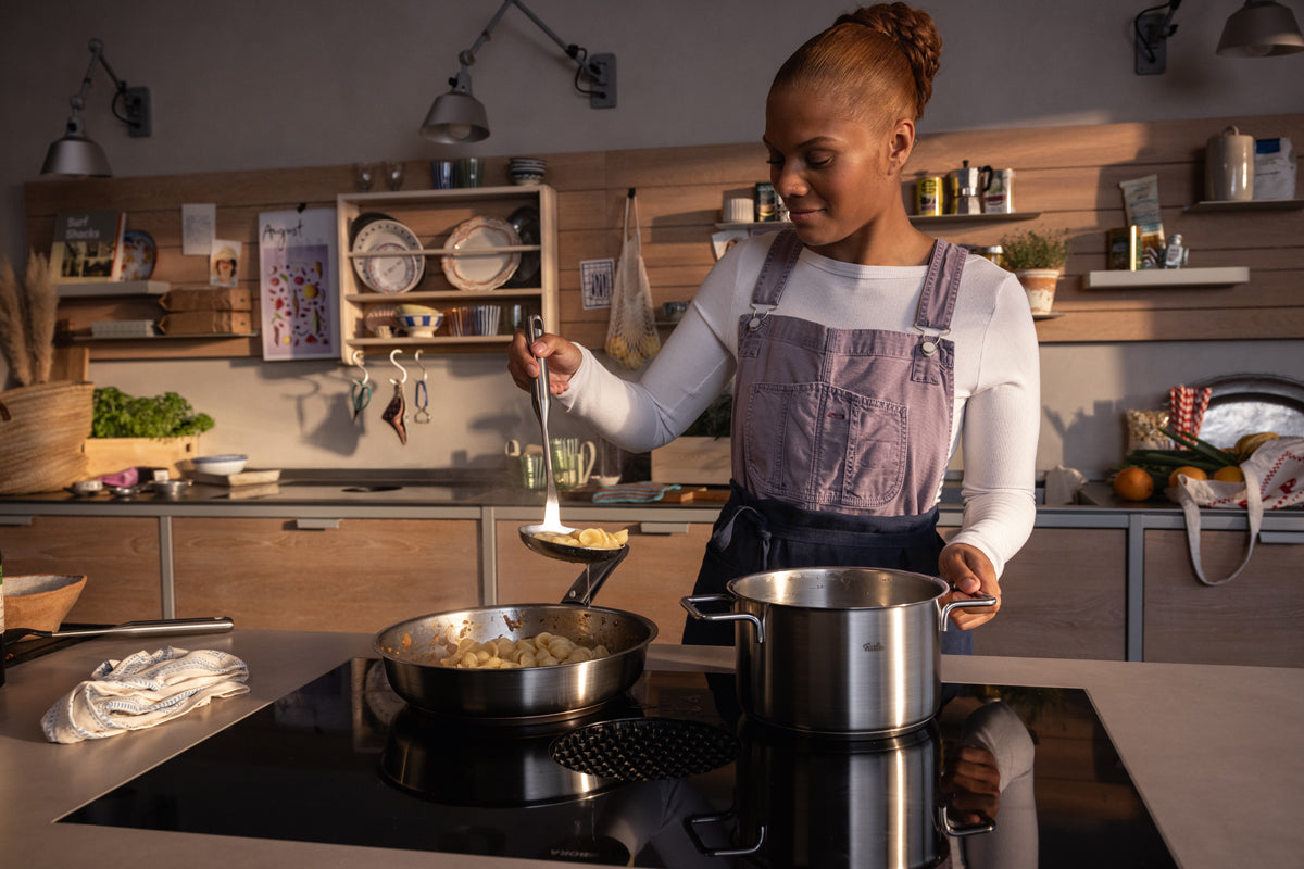 A woman smiles in a modern kitchen and cooks with the Fissler Pure Collection Cooking Pot with Metal Lid, known for quality Made in Germany.