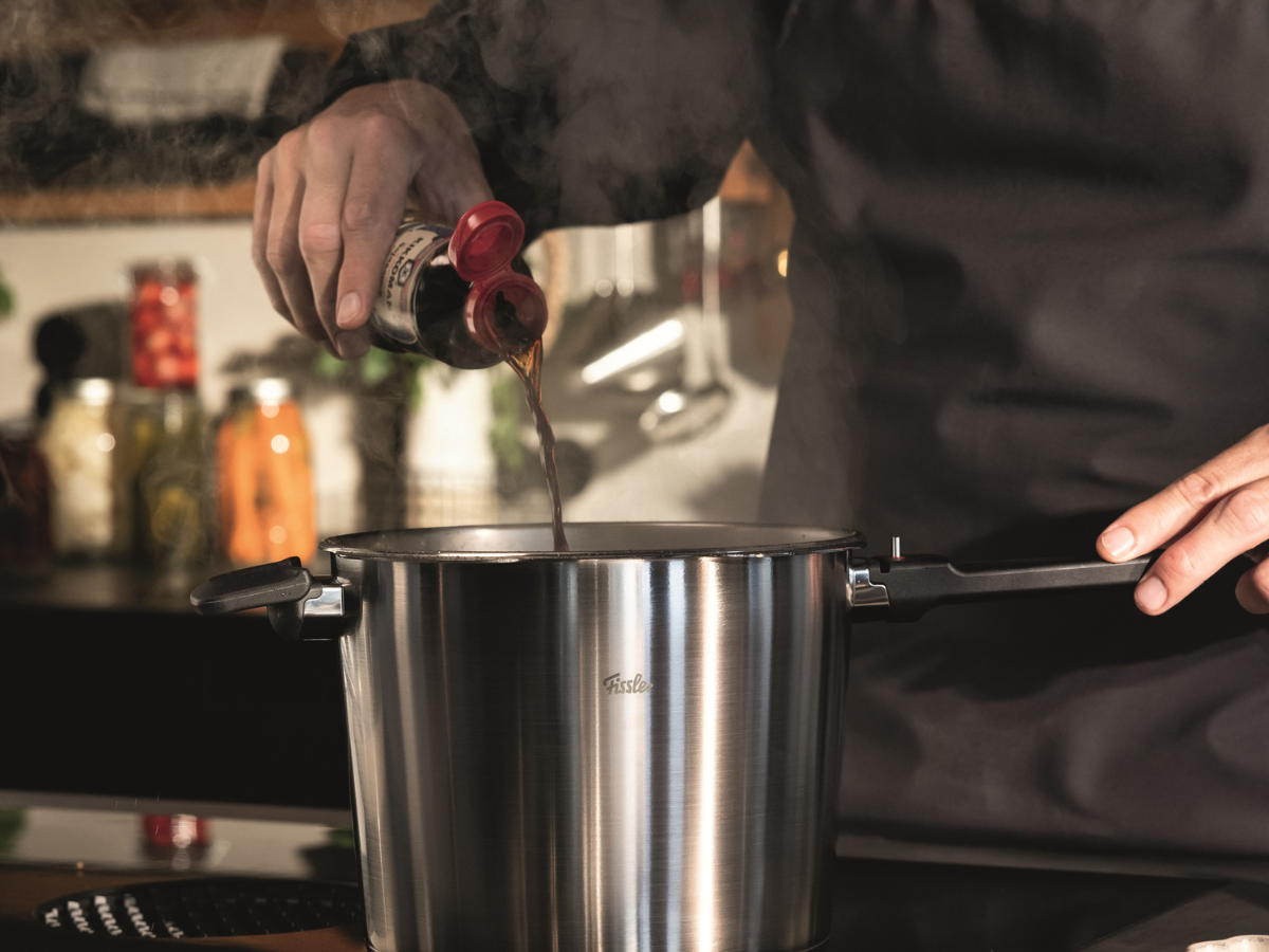 Eine Person in einer dunklen Schürze gießt Soße aus einer Flasche in einen dampfenden Fissler-Edelstahltopf auf einem Herd in einer modernen Küche und verdeutlicht damit das Bekenntnis der Marke zu präziser Technik, Langlebigkeit und zeitlosem deutschem Design.