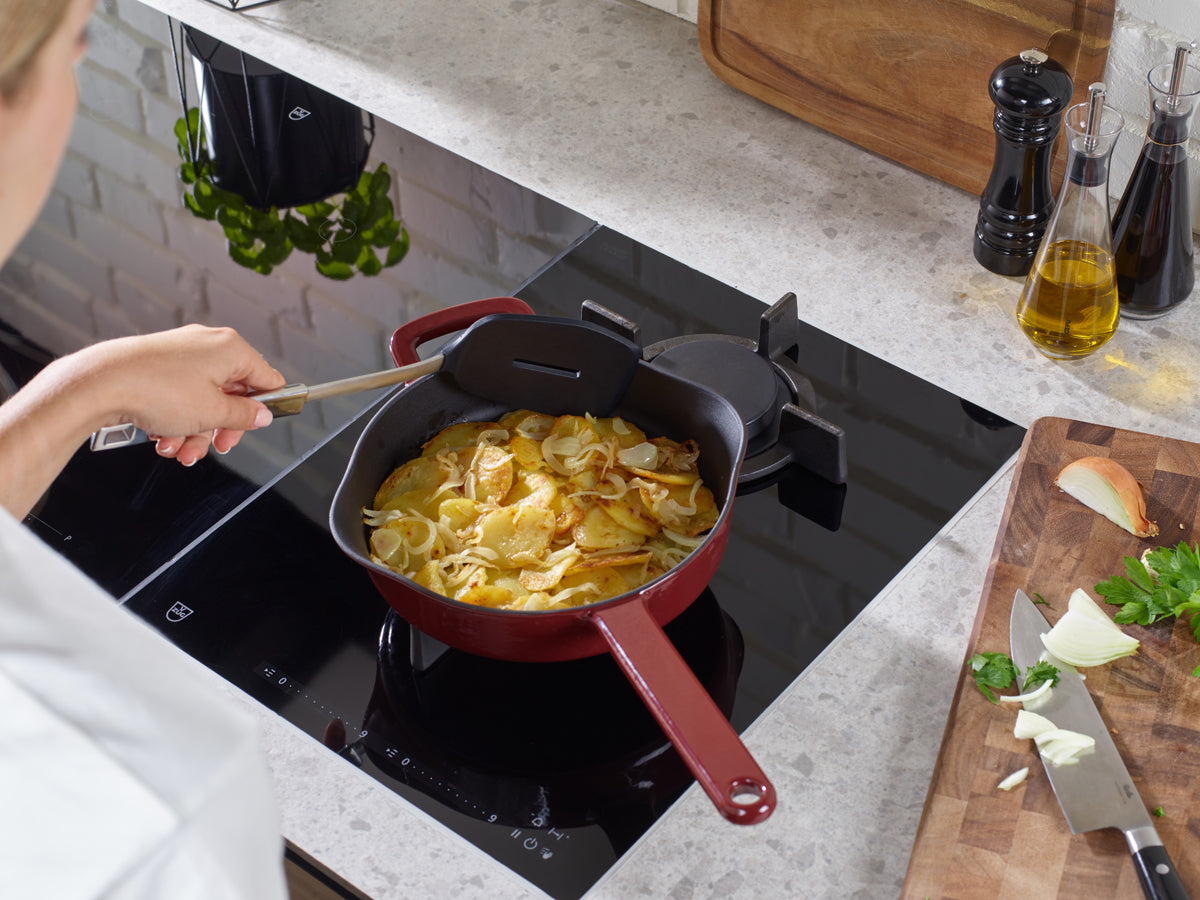 Eine Person rührt Kartoffeln und Zwiebeln in einer roten Fissler Gusseisen Pfanne; frische Zutaten und Streuer vervollständigen diese moderne Küchenszene.