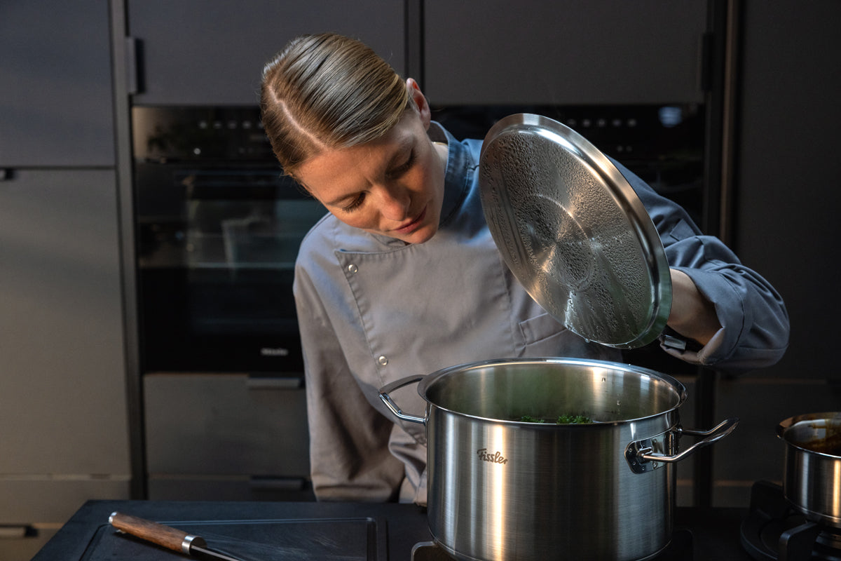 Ein Koch in grauer Uniform hebt den Deckel eines Fissler-Edelstahltopfes auf dem Herd an und zeigt damit deutsche Technik in einer modernen Küche.