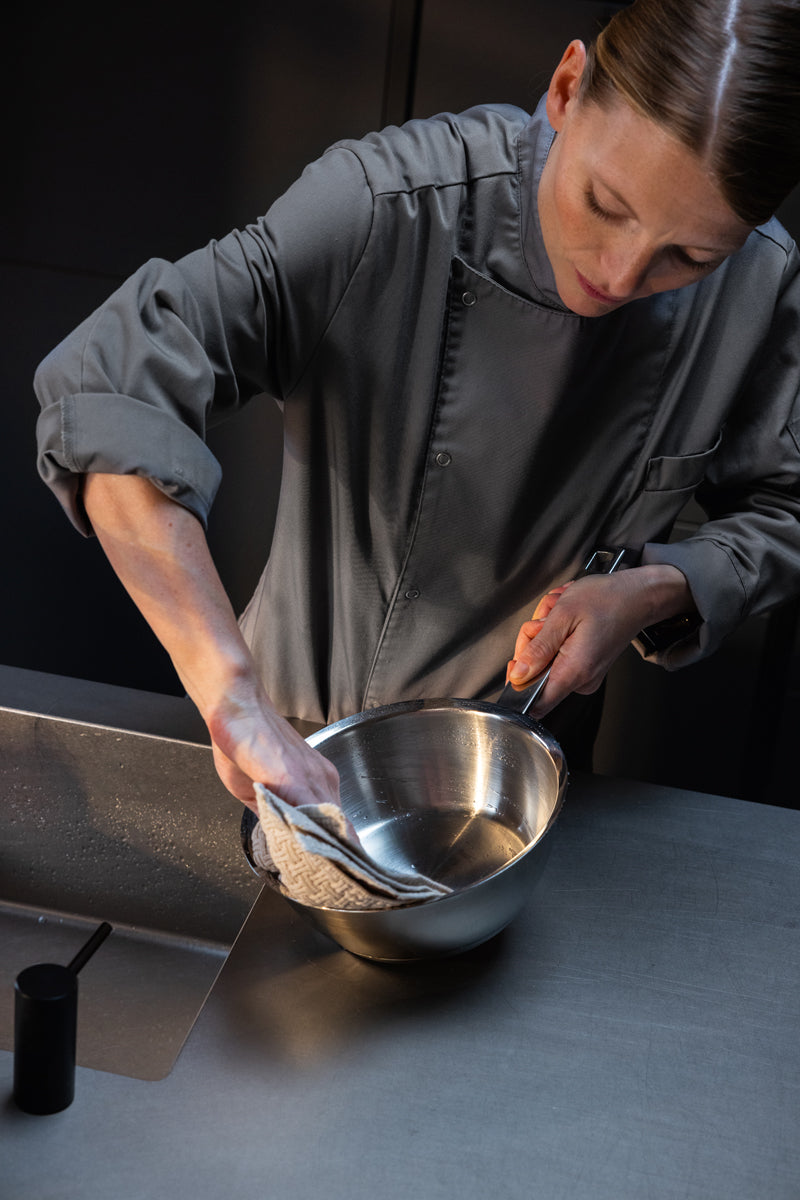 Ein Koch in grauer Uniform wischt eine Sauteuse (20 cm) aus der Fissler Original-Profi Collection® in einem Spülbecken einer Großküche mit einem Tuch ab – ein Beispiel für Fisslers Tradition präzisionsgefertigter, langlebiger Kochgeschirrprodukte aus deutscher Herstellung.