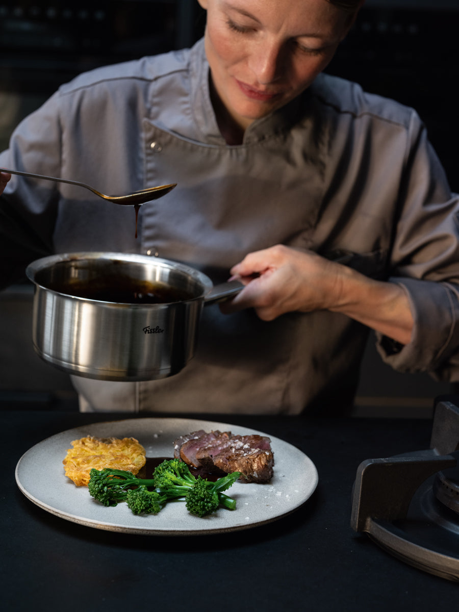 Ein Koch in grauer Uniform gießt gekonnt mit Hilfe des 7-teiligen Edelstahltopf-Sets „Fissler Original-Profi Collection®“ mit Metalldeckeln Soße aus einem unserer präzise gefertigten Töpfe auf ein wunderschön angerichtetes Steak, Broccolini und einen Kartoffelkuchen. Diese elegante Anrichtung unterstreicht die professionelle Qualität und das zeitlose Design des Fissler-Kochgeschirrs – seit 1845 in Deutschland gefertigt, um Langlebigkeit und Innovation in jeder Küche zu gewährleisten.