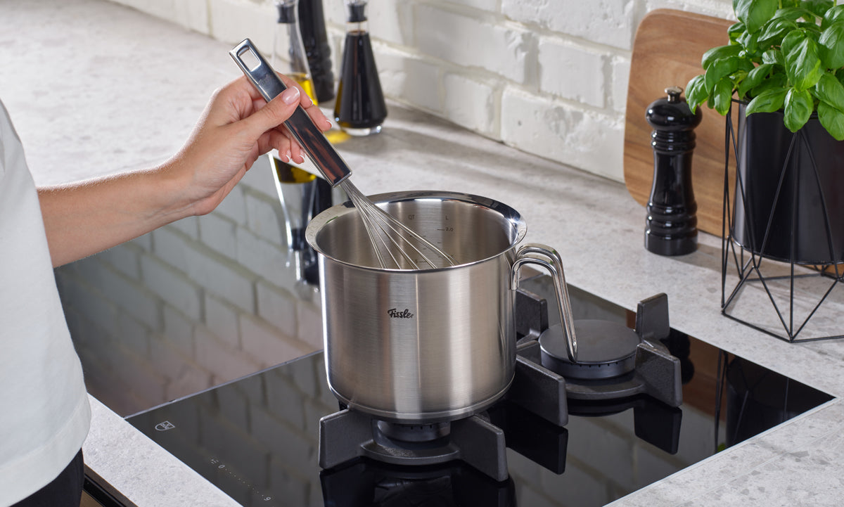 Eine Person rührt mit einem Schneebesen in einem Milchtopf (16 cm) aus der Fissler Original-Profi Collection®, der dank präziser Fertigung besonders langlebig ist, auf einem Gasherd um.