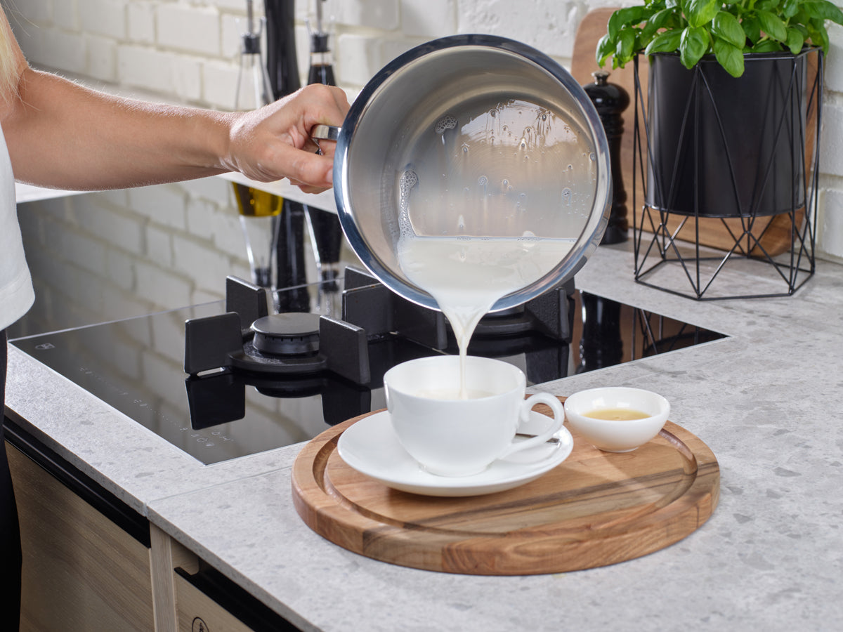 Eine Hand gießt Milch aus einem 16 cm großen Milchkännchen der Fissler Original-Profi Collection® – das sich durch deutsche Präzision auszeichnet – in eine weiße Teetasse, die auf einem Holzbrett steht.