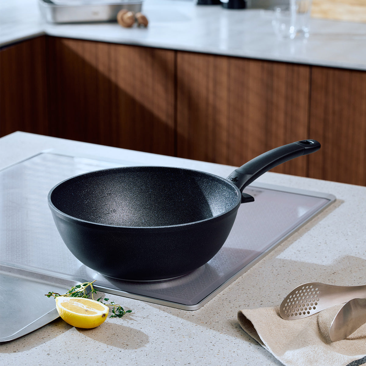 Der Fissler Adamant® Wok mit seinem robusten Griff und der eleganten schwarzen Oberfläche steht stilvoll auf einer Marmorarbeitsplatte, während im Hintergrund die Wärme der Holzschränke zu spüren ist. Dieser Wok, der seit 1845 in Deutschland hergestellt wird, verkörpert Fisslers Tradition von Langlebigkeit und Innovation durch seine fortschrittliche, kratzfeste Antihaftbeschichtung – perfekt für kulinarische Kreationen im Alltag. Abgerundet wird das Bild durch frische Kräuter, eine in Scheiben geschnittene