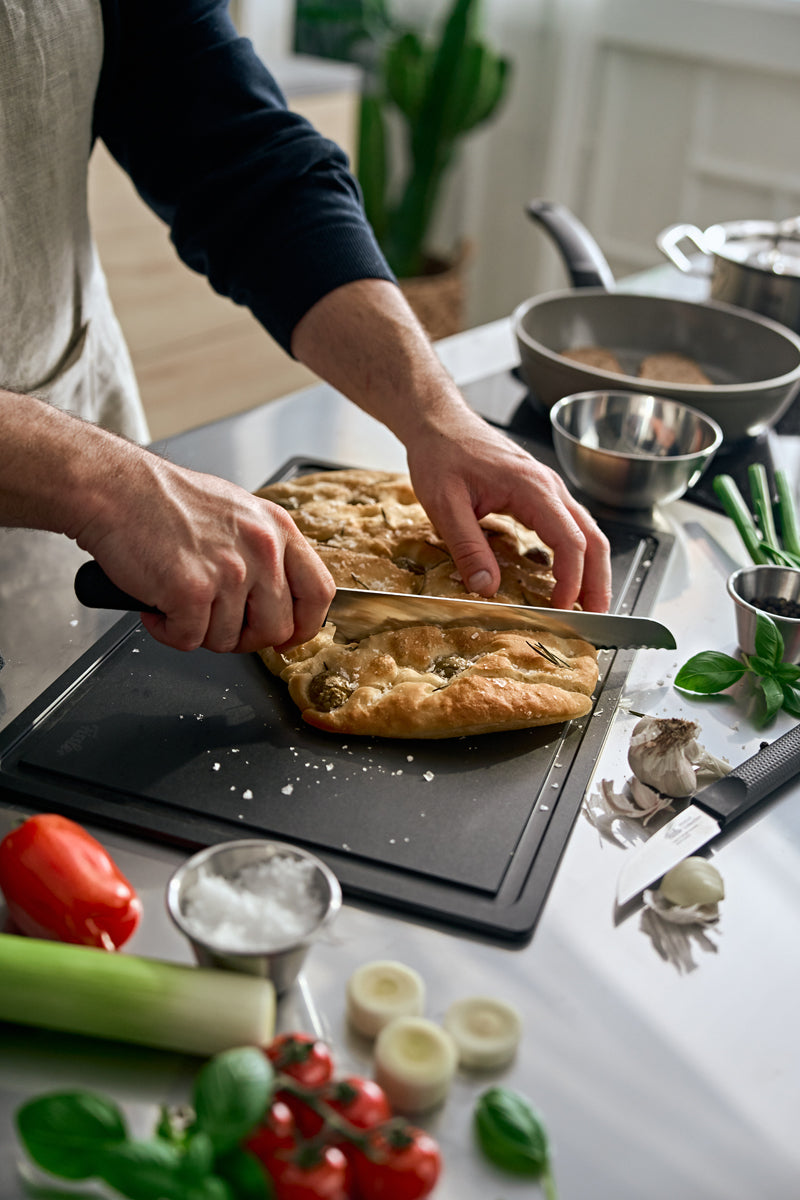 Eine Person schneidet frisches Brot mit dem präzisionsgefertigten Fissler ProCut Brotmesser 21 cm, das über eine extraharte Stahl-Wellenschliffklinge verfügt, auf einem Schneidebrett, umgeben von frischem Gemüse, Kräutern, Salz und hochwertigen Küchenutensilien in einer hellen, modernen Küche – eine Kulisse, die Fisslers Engagement für zeitloses Design, Langlebigkeit und Innovation unterstreicht, das unser Kochgeschirr „Made in Germany“ auszeichnet.