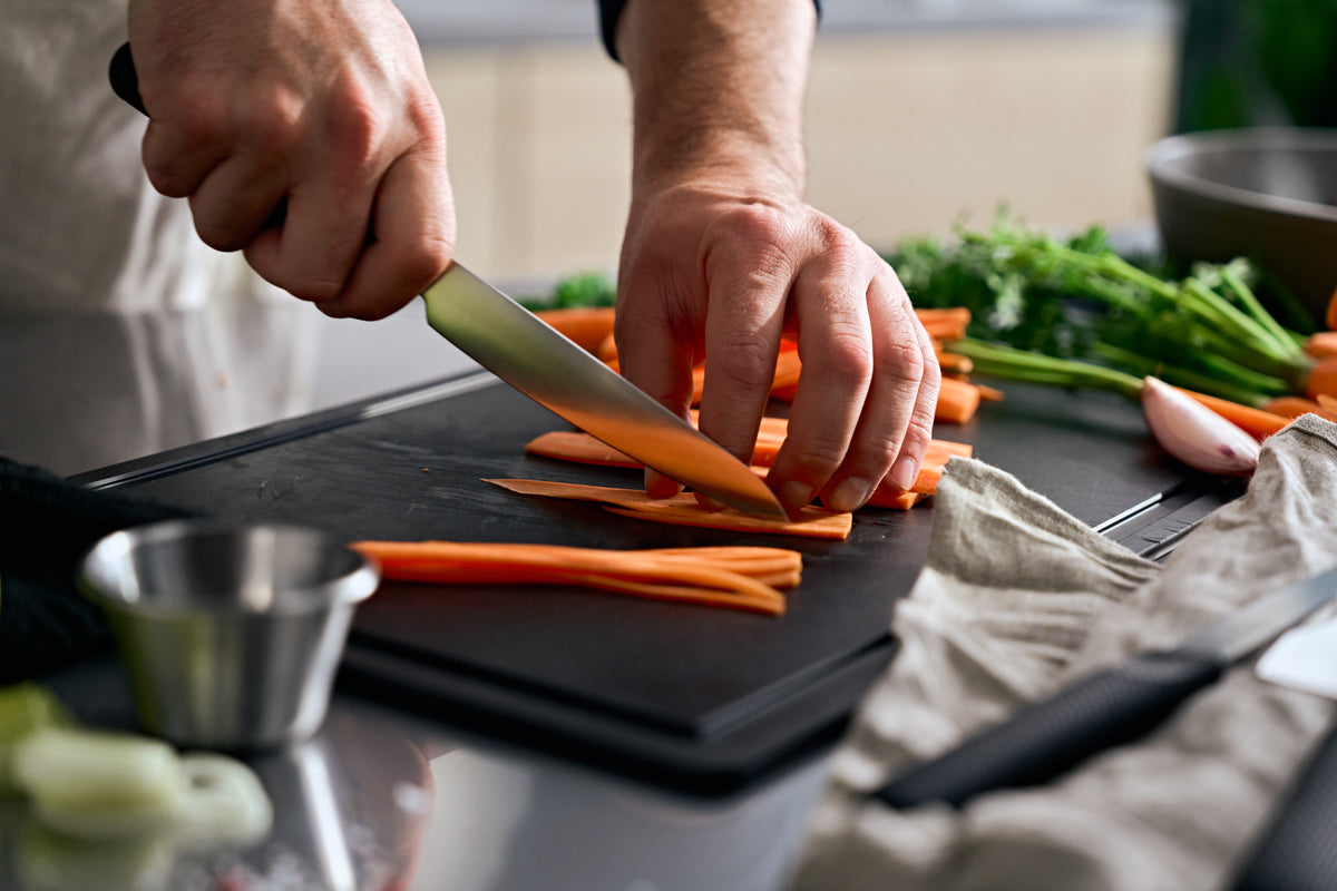 In einer modernen Küche schneidet jemand gekonnt Möhren auf einem eleganten schwarzen Schneidebrett mit dem Fissler ProCut Universalmesser 14 cm – einem präzisionsgefertigten deutschen Messer, das die langjährige Tradition von Fissler in Sachen Innovation und Qualität seit 1845 widerspiegelt. Umgeben von frischem Gemüse und hochwertigem Fissler-Kochzubehör unterstreicht diese Szene das zeitlose Design und die Langlebigkeit, die den Kern jedes Fissler-Produkts „Made in Germany“ ausmachen.