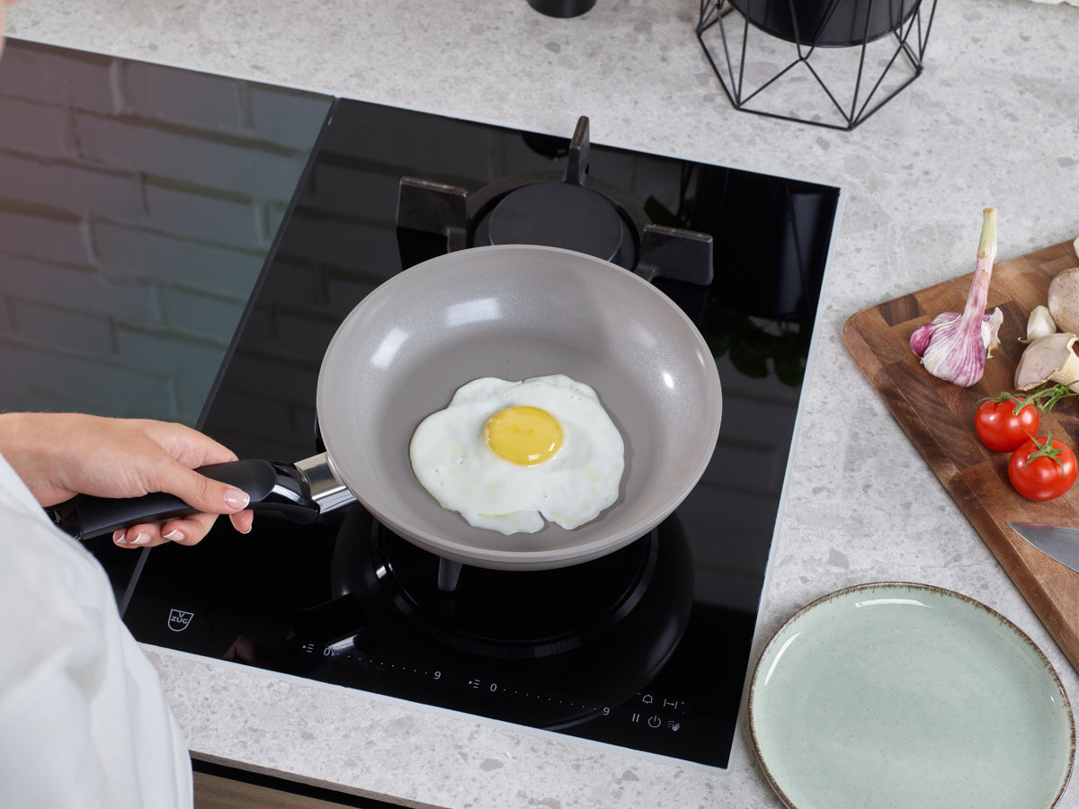 Eine Person bereitet auf dem Herd ein Spiegelei im Fissler 2-tlg. Pfannen-Set Ceratal® Classic 28 cm zu, einer hochwertigen, in Deutschland hergestellten Pfanne mit PFAS-freier Keramikbeschichtung. Neben der Pfanne liegen auf einem Schneidebrett frische Tomaten, Knoblauch und Pilze sowie ein leerer Teller – ein Zeichen für Fisslers Engagement für präzise Handwerkskunst und langlebiges Kochgeschirr, das für zeitlose Küchen geschaffen wurde.