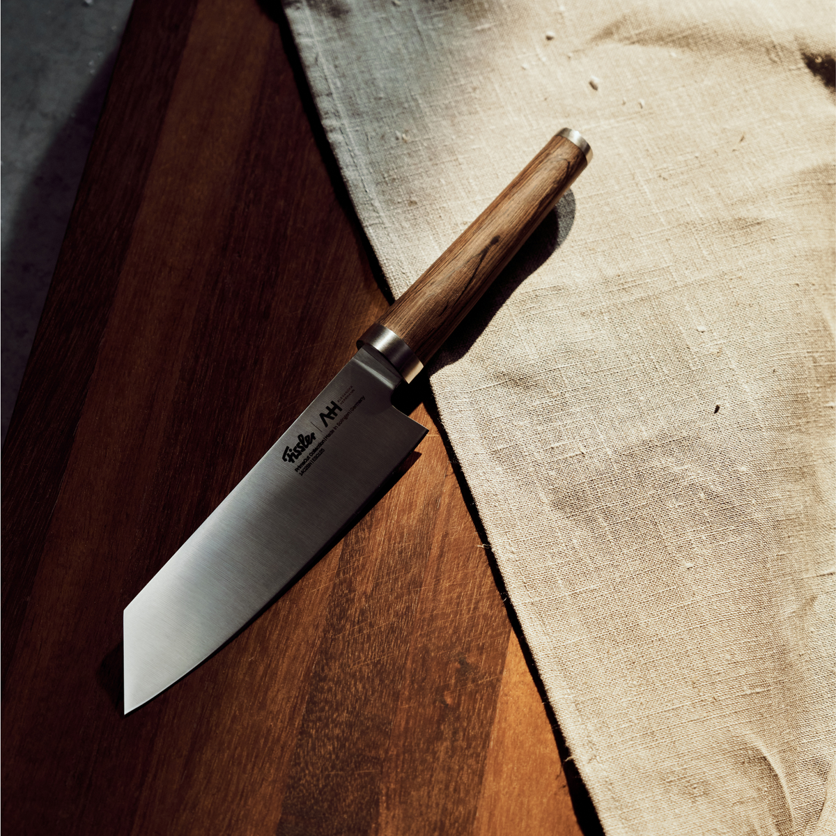 Ein Fissler Kochmesser mit einem handgefertigten Holzgriff auf Holz neben einem beigen Leinen, das präzises deutsches Design in dramatischem Licht präsentiert.