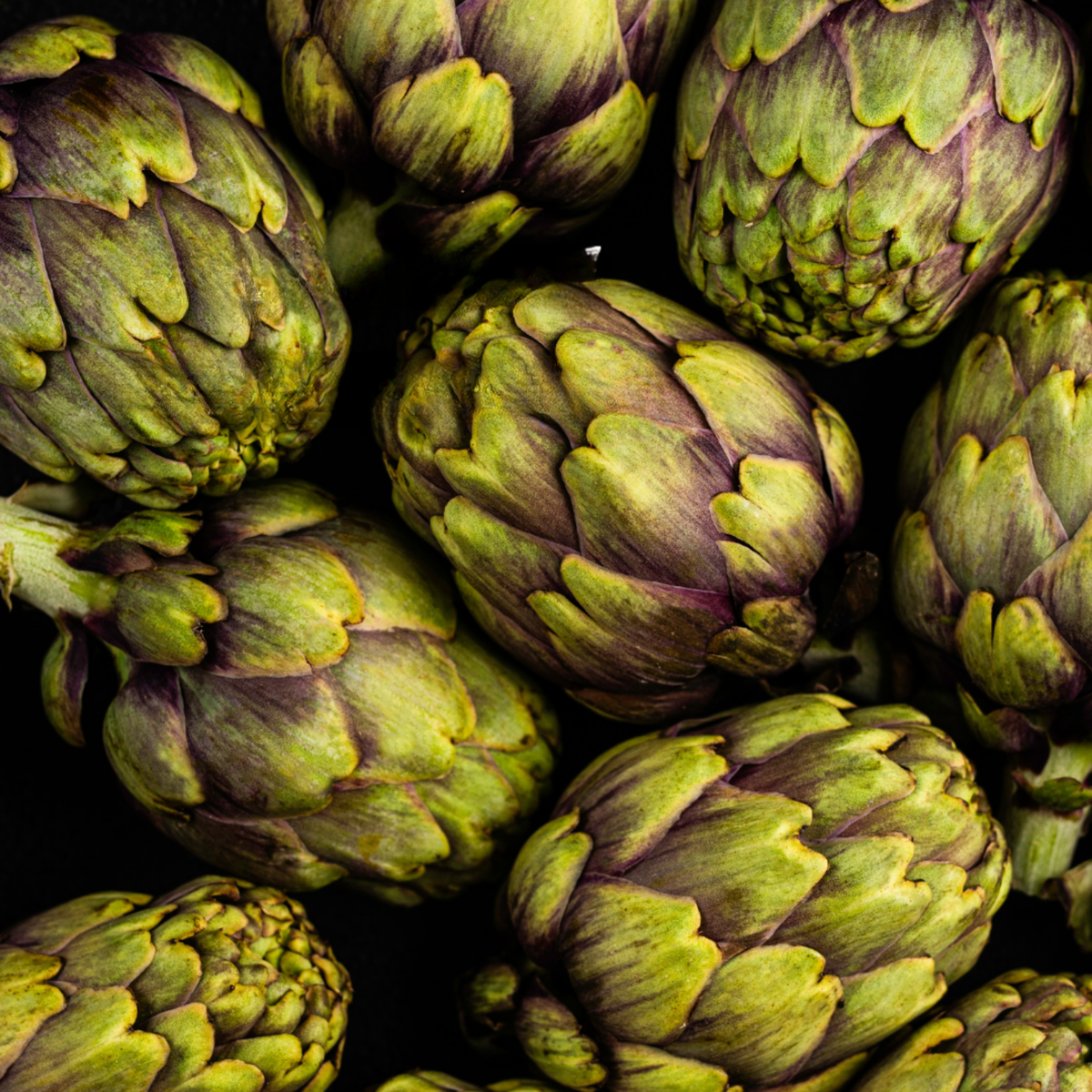 Frische Artischocken mit grünen und violetten Farbtönen, die in einer Fissler-Edelstahlpfanne vor einem dunklen Hintergrund angeordnet sind.