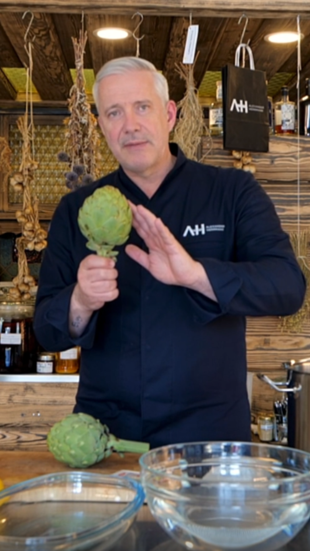 Ein Koch in einer rustikalen Küche hält eine frische Artischocke neben Fissler-Kochgeschirr und Glasschüsseln; im Hintergrund hängen zeitlose Werkzeuge.