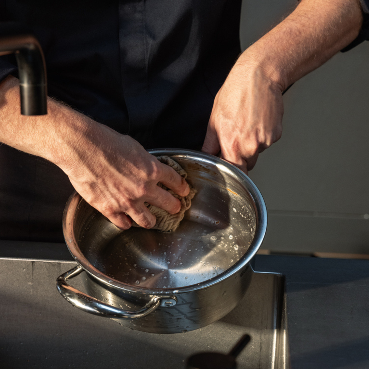 Eine Person reinigt einen Fissler-Edelstahltopf mit einem Schwamm am Spülbecken und demonstriert damit, wie pflegeleicht unser präzise gefertigtes Kochgeschirr aus deutscher Produktion ist. Das elegante, langlebige Design besticht auch bei der täglichen Pflege.