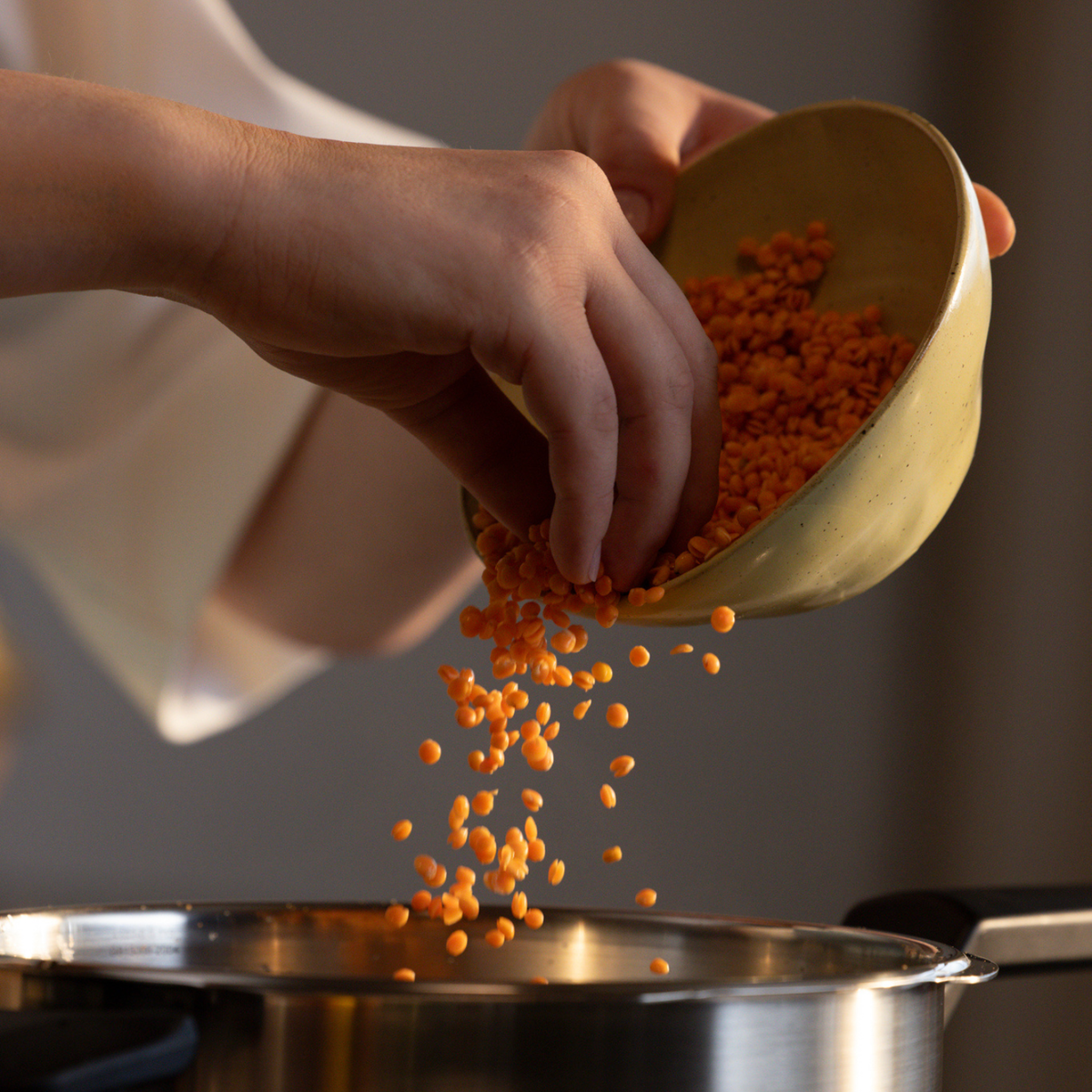 Eine Person schüttet rote Linsen aus einer gelben Schüssel in einen hochwertigen Fissler-Edelstahltopf auf einem Herd und unterstreicht damit die Langlebigkeit und das zeitlose Design des in Deutschland entwickelten Kochgeschirrs von Fissler.