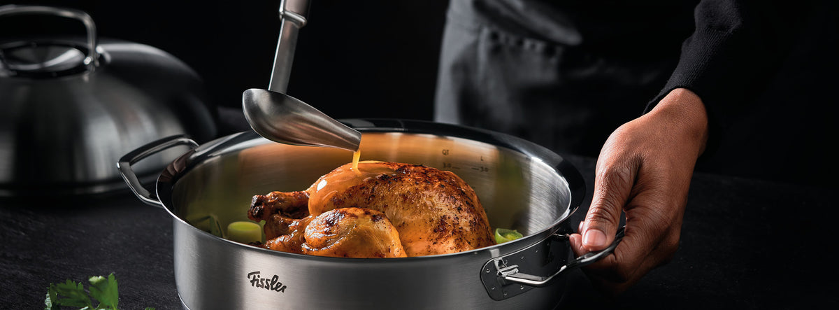 Die Soße wird über das Brathähnchen in einem Fissler-Edelstahltopf gegossen, mit Kräutern und Deckel in der Nähe - ein Beispiel für deutsche Qualitätskochgeschirre.