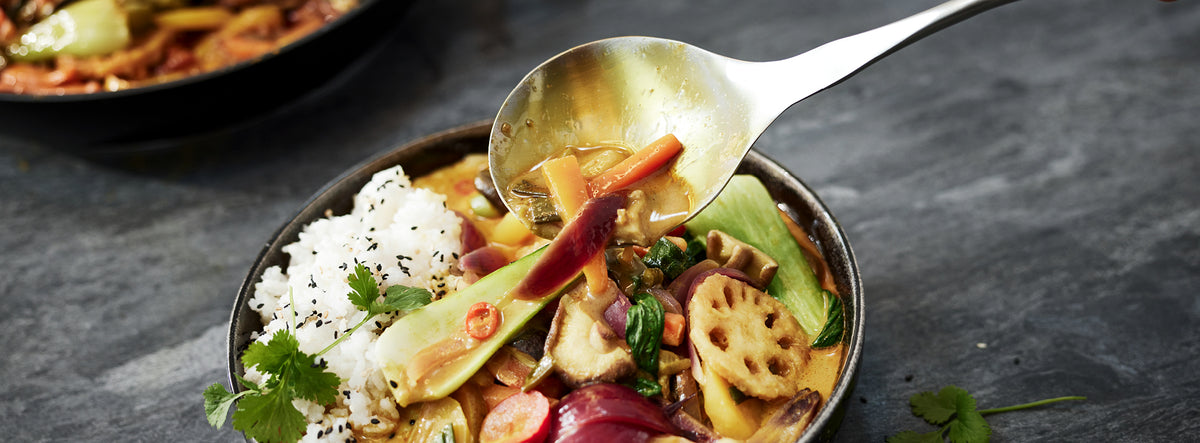 Ein lebhaftes Gemüsecurry mit Reis, serviert in einer Fissler-Schüssel. Ein Löffel hebt das Curry auf und zeigt die Exzellenz des deutschen Kochgeschirrs.