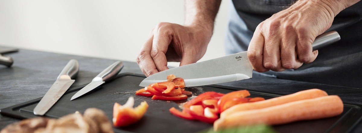Eine Person schneidet eine rote Paprika auf einem Fissler-Schneidebrett mit einem hochwertigen Fissler-Kochmesser, daneben liegen Karotten und Messer.