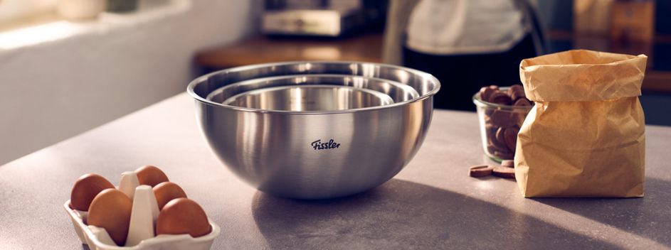 Drei präzise gefertigte Fissler-Rührschüsseln aus Edelstahl, die elegant auf einer Küchenarbeitsplatte ineinander gestapelt sind, stehen beispielhaft für zeitlose deutsche Handwerkskunst. Daneben unterstreichen hochwertige Backzutaten – eine Papiertüte mit Mehl, eine Glasschüssel mit Schokoladenstückchen und ein Karton mit Eiern – die kulinarische Kreativität. Im Hintergrund steht eine Person, bereit, ihre Inspiration mit der bekannten Langlebigkeit und dem innovativen Design von Fissler zum Leben zu