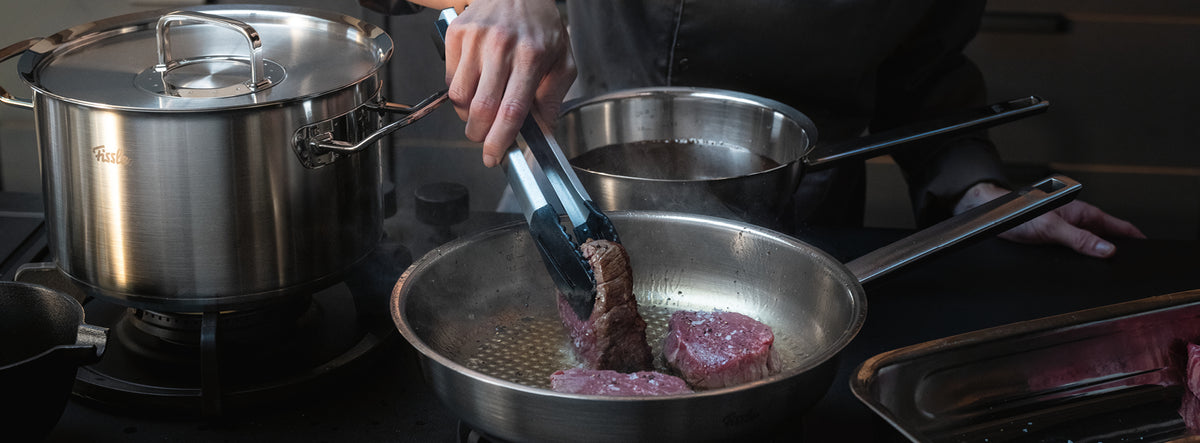 Eine Person brät Fleisch mit einer Zange in einer Fissler-Edelstahlbratpfanne an, die neben unserem charakteristischen Topf und der Kasserolle auf dem Herd steht.