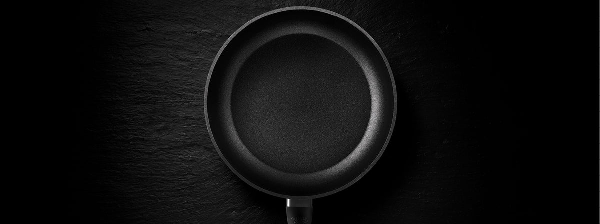 Eine schwarze antihaftbeschichtete Bratpfanne von Fissler, von oben gesehen, auf einer dunkel strukturierten Oberfläche - präzise Technik trifft auf zeitloses Design.