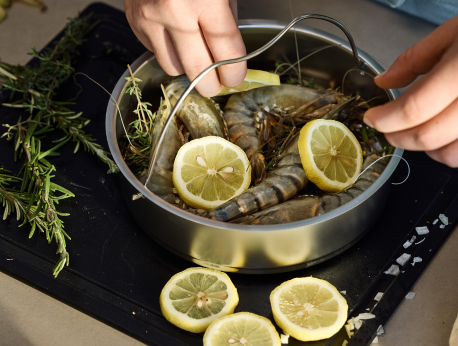 Eine Person bereitet rohe Garnelen in einem hochwertigen Fissler-Edelstahltopf zu, gekonnt garniert mit frischen Kräutern und Zitronenscheiben. Weitere Zitronen und Kräuter liegen auf einem Schneidebrett daneben und unterstreichen die Präzision und das zeitlose Design der in Deutschland entwickelten Fissler-Kochgeschirrserie.