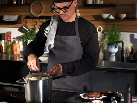 Eine Person, die eine Brille, eine Mütze und eine Schürze trägt, hebt in einer modernen Küche den Deckel eines Fissler-Edelstahltopfs ab. Auf der Arbeitsplatte daneben liegen gegartes Fleisch und hochwertige Küchenutensilien, im Hintergrund sind in Regalen weiteres Kochgeschirr und Pflanzen zu sehen – ein Beleg für Fisslers Engagement für Präzisionstechnik, Langlebigkeit und zeitloses deutsches Design.