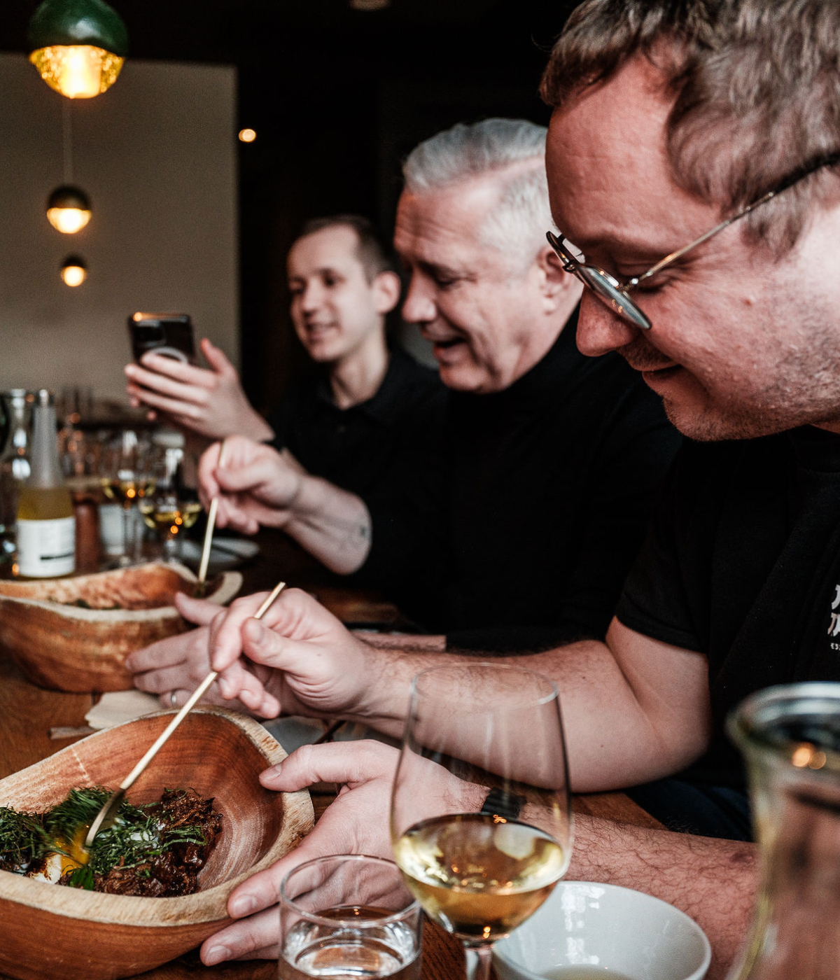 Drei Menschen genießen gemeinsam eine Mahlzeit am Tisch, lächeln und lachen miteinander. Frisch zubereitete Gerichte werden in Holzschalen serviert, daneben stehen Getränke. Hochwertiges Fissler-Edelstahlkochgeschirr und elegante Küchenaccessoires runden das Bild ab und spiegeln deutsche Handwerkskunst und zeitloses Design wider. Eine Person hält ein Smartphone in der Hand, während eine andere mit Essstäbchen isst – ein Beispiel für modernes Essverhalten in Kombination mit den präzise gefertigten