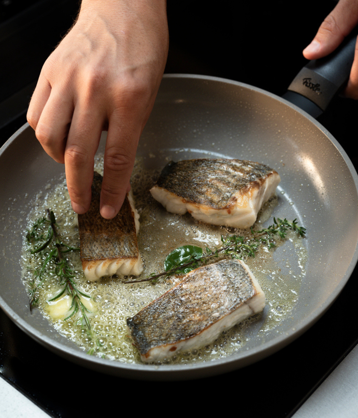 Drei Fischstücke mit Haut werden in einer Fissler-Pfanne fachmännisch angebraten, umgeben von sprudelnder Butter, frischen Kräutern und Knoblauch – ein Beispiel für präzises Kochen mit hochwertigem Kochgeschirr aus deutscher Fertigung.