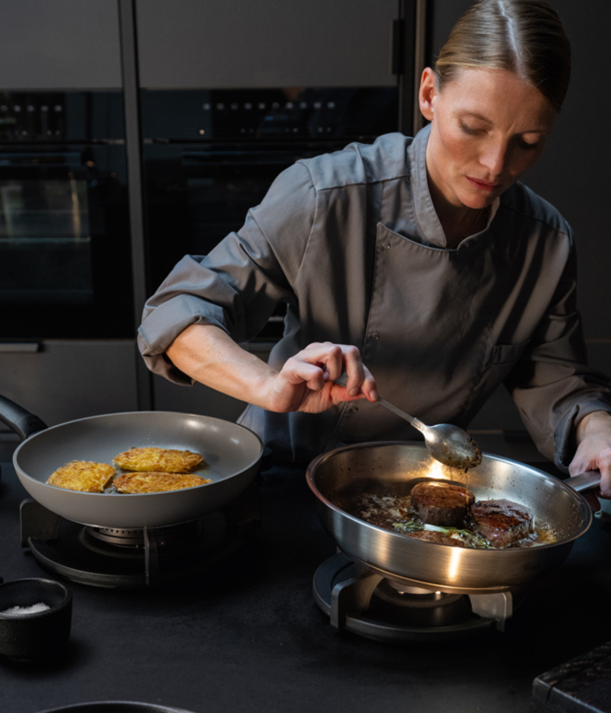Eine Köchin in grauer Uniform bereitet Fleisch mit größter Präzision zu und begießt es dabei in einer hochwertigen Fissler-Edelstahlpfanne auf dem Herd. Neben ihr stehen in einer weiteren Fissler-Pfanne goldbraune Kartoffelpuffer – alles in einer eleganten, modernen Küche, die das Bekenntnis der Marke zu deutscher Ingenieurskunst und zeitlosem Design unterstreicht.