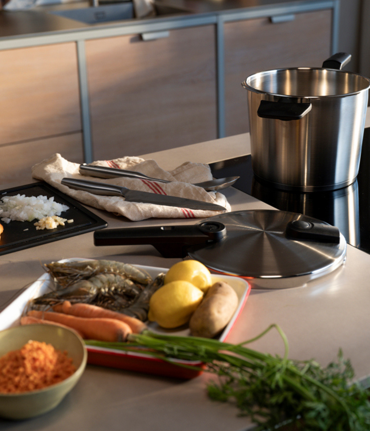 Auf einer Küchenarbeitsplatte steht ein Fissler-Edelstahltopf mit passendem Deckel, umgeben von frischen Möhren, Zitronen, Kartoffeln, gehackten Zwiebeln und einer Schüssel mit roten Linsen. Hochwertige Fissler-Kochutensilien sind ordentlich auf einem Handtuch daneben angeordnet und unterstreichen die Präzision deutscher Ingenieurskunst sowie das zeitlose Design.