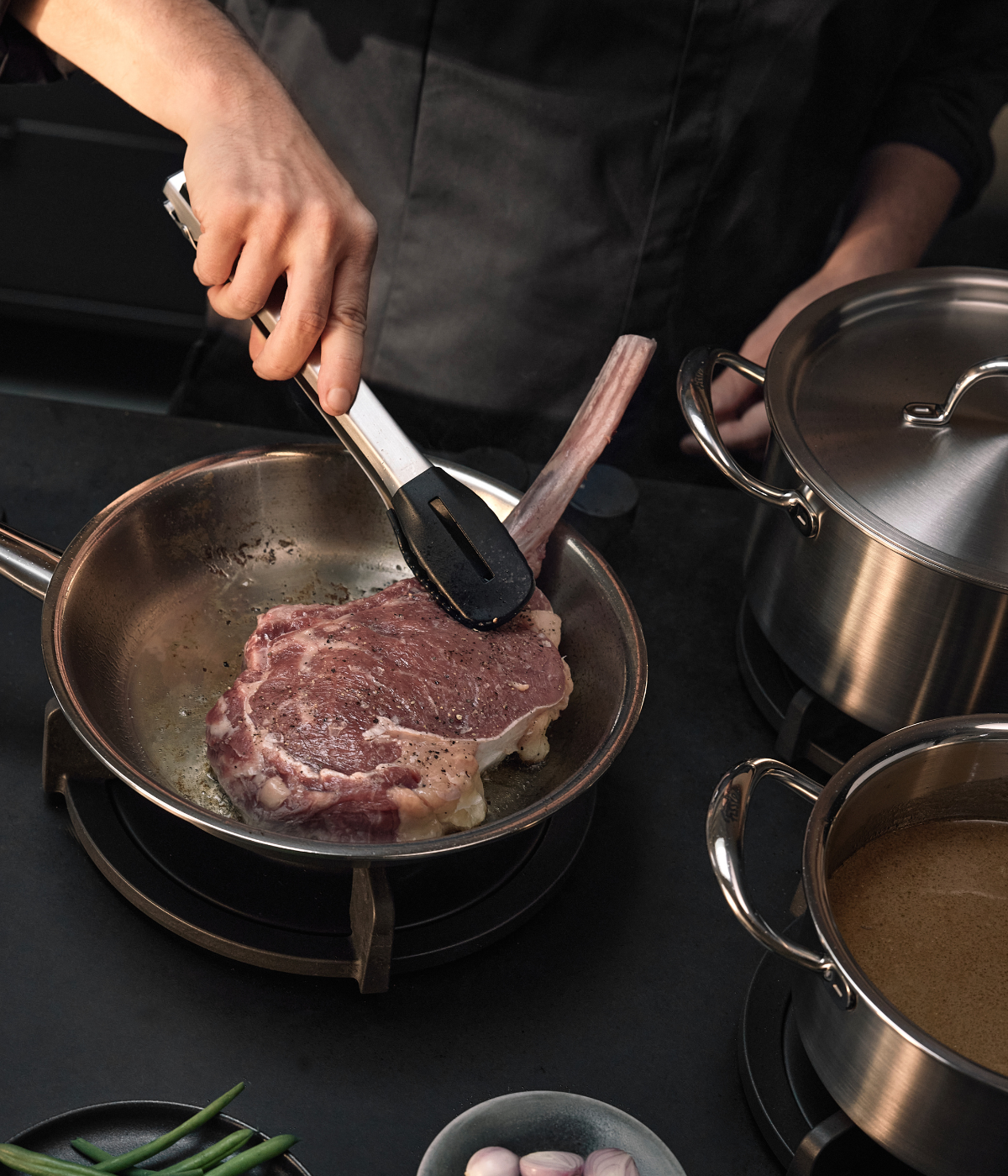 Eine Person brät gekonnt ein großes Steak mit Knochen in einer hochwertigen Fissler-Edelstahlpfanne mit Hilfe einer Zange an. Daneben stehen zwei Fissler-Kochtöpfe auf dem Herd, die sich durch Präzision und Langlebigkeit auszeichnen, während im unteren Bildbereich frische grüne Bohnen und Schalotten zu sehen sind – ein Bild, das eine Küchenszene geprägt von deutscher Ingenieurskunst und zeitlosem Design unterstreicht.