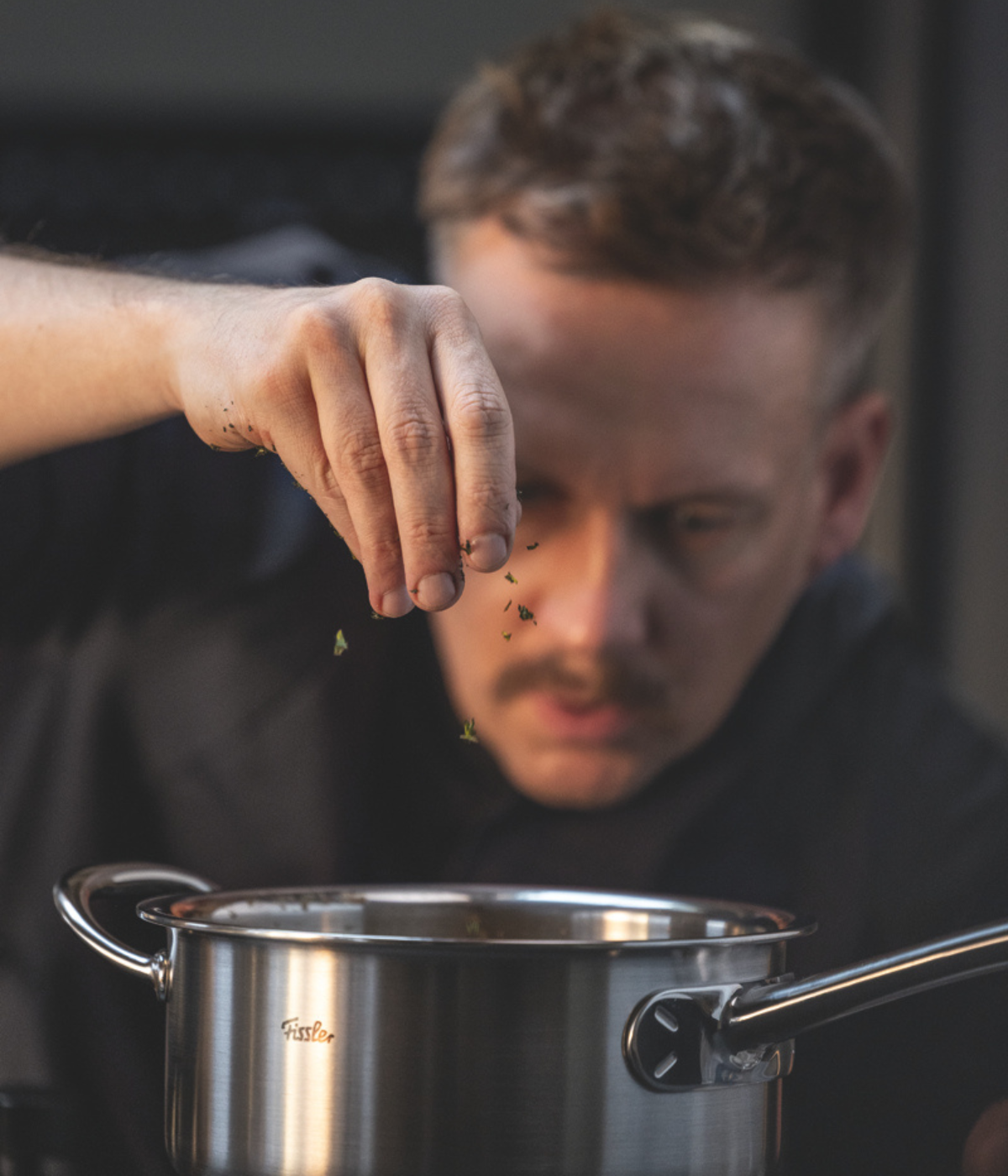 Ein Koch gibt frische Kräuter in einen Fissler-Edelstahltopf und demonstriert dabei Präzision und Konzentration, während die elegante deutsche Ingenieurskunst und das zeitlose Design des Topfes in der Küchenszene deutlich zum Ausdruck kommen.