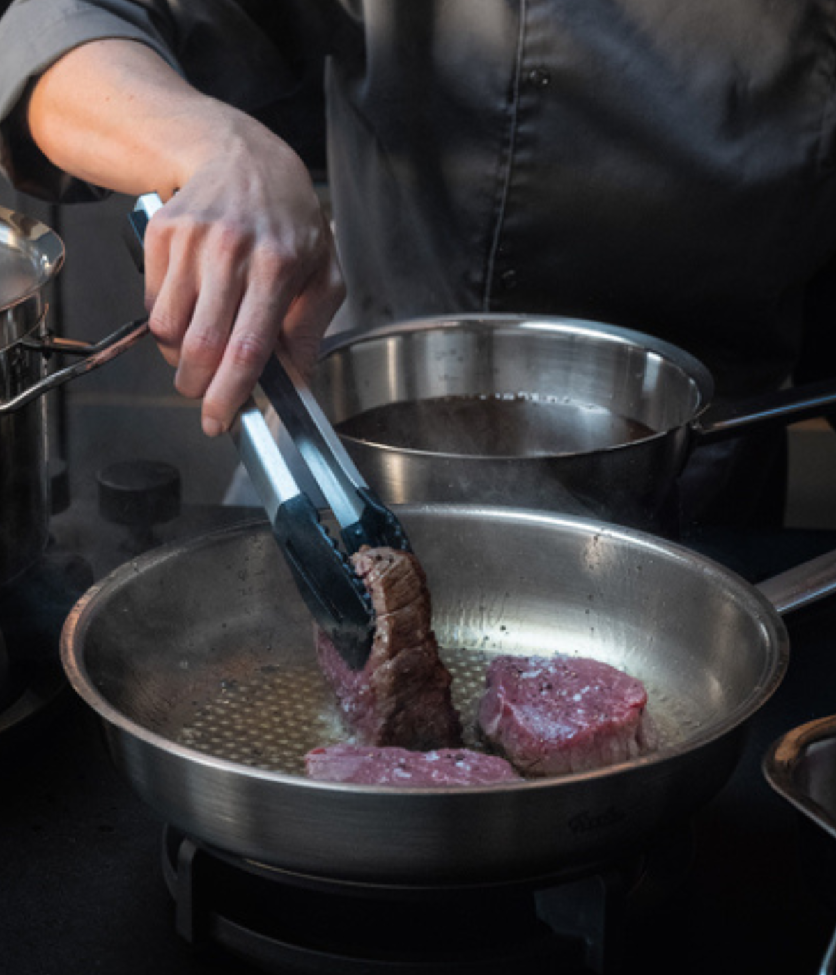 Ein Koch in einer dunklen Jacke brät Steakstücke gekonnt mit einer Zange in einer Fissler-Edelstahlpfanne auf dem Herd an und unterstreicht damit die präzise Technik und das zeitlose deutsche Design.
