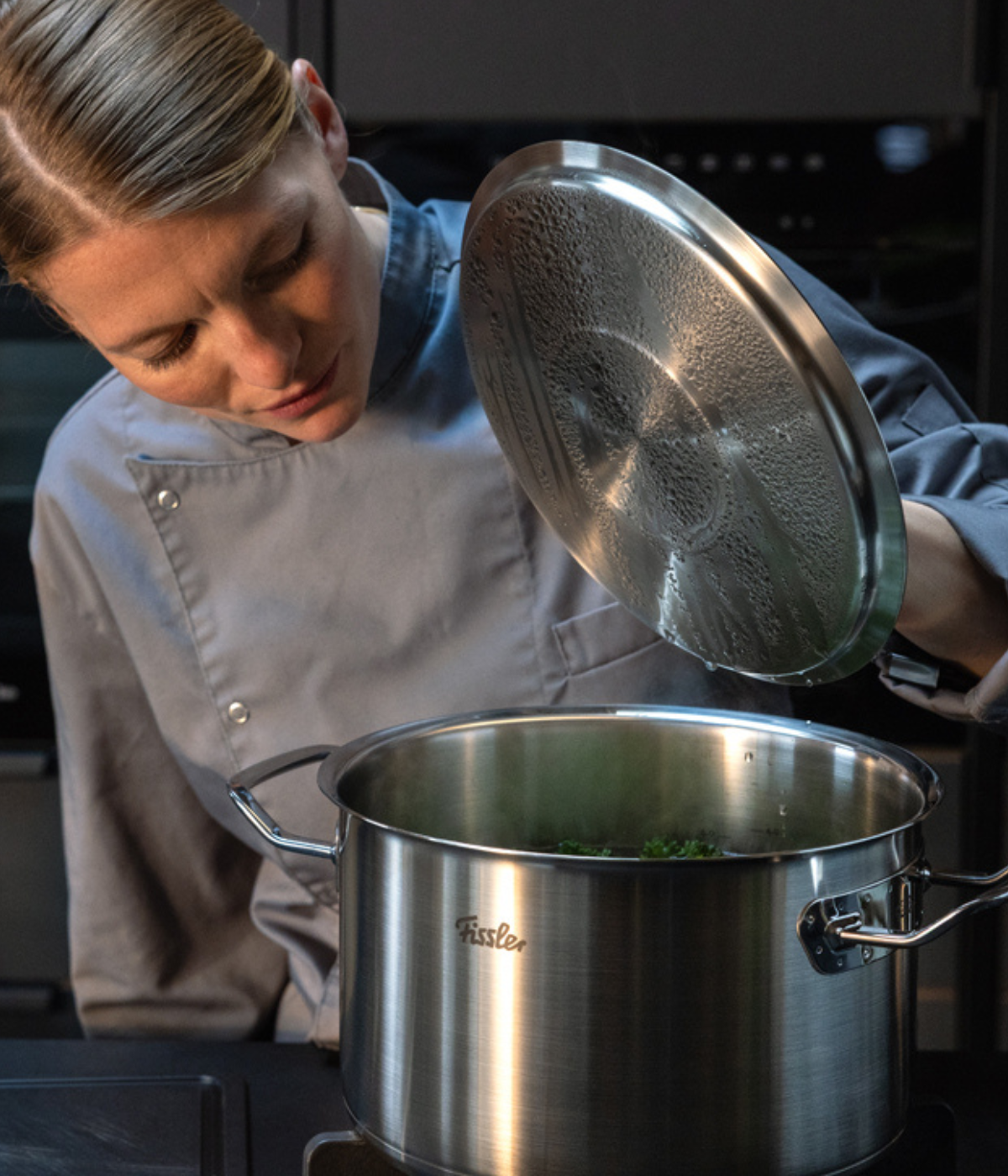 A chef in a gray uniform lifts the lid of a premium Fissler stainless steel pot on a stove, showcasing precision engineering and timeless German design while checking the ingredients inside.