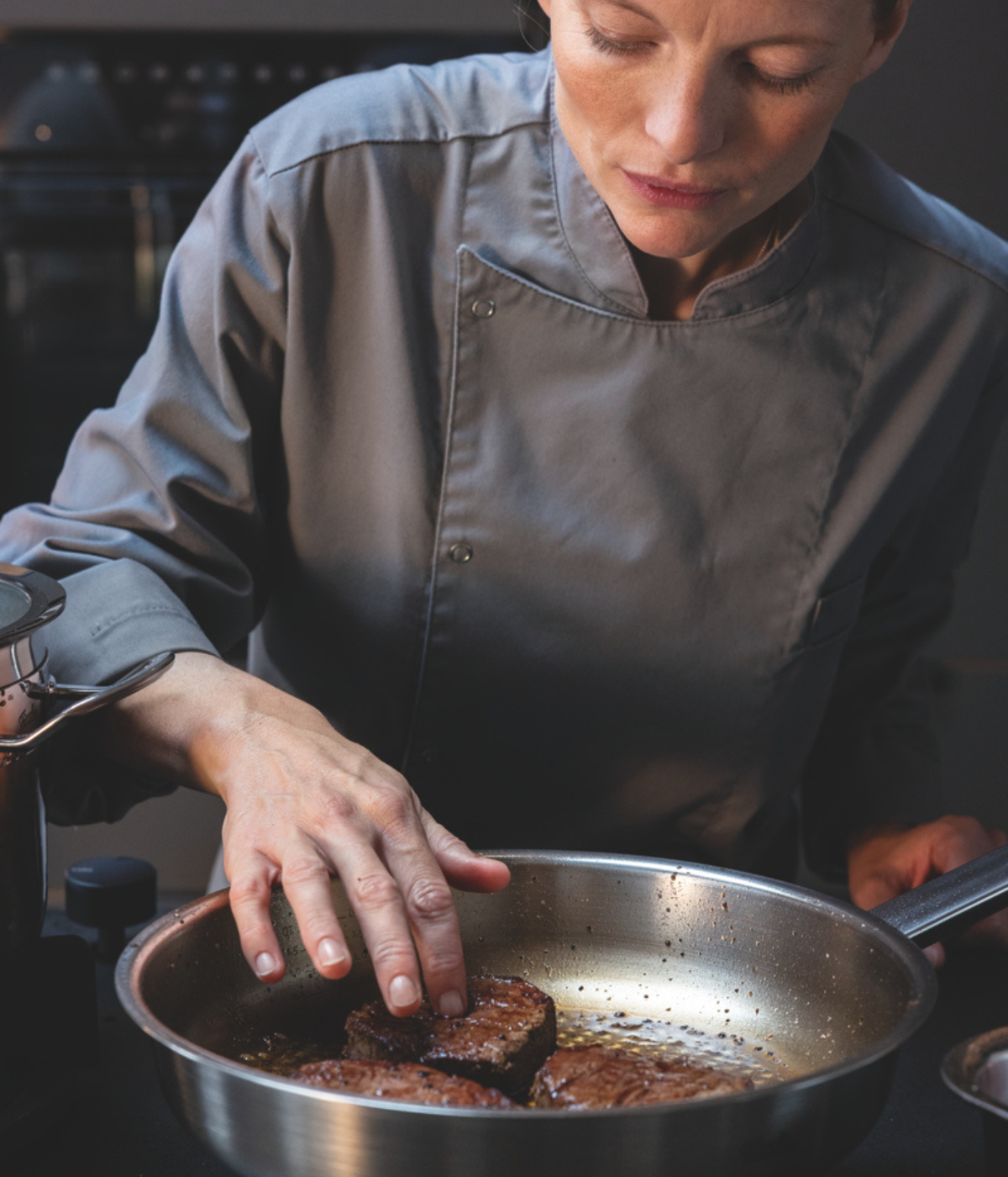 Ein Koch in grauer Uniform prüft den Gargrad zweier Steaks, die in einer Fissler-Edelstahlpfanne brutzeln – ein Beispiel für Präzision und hochwertige Technik „Made in Germany“.