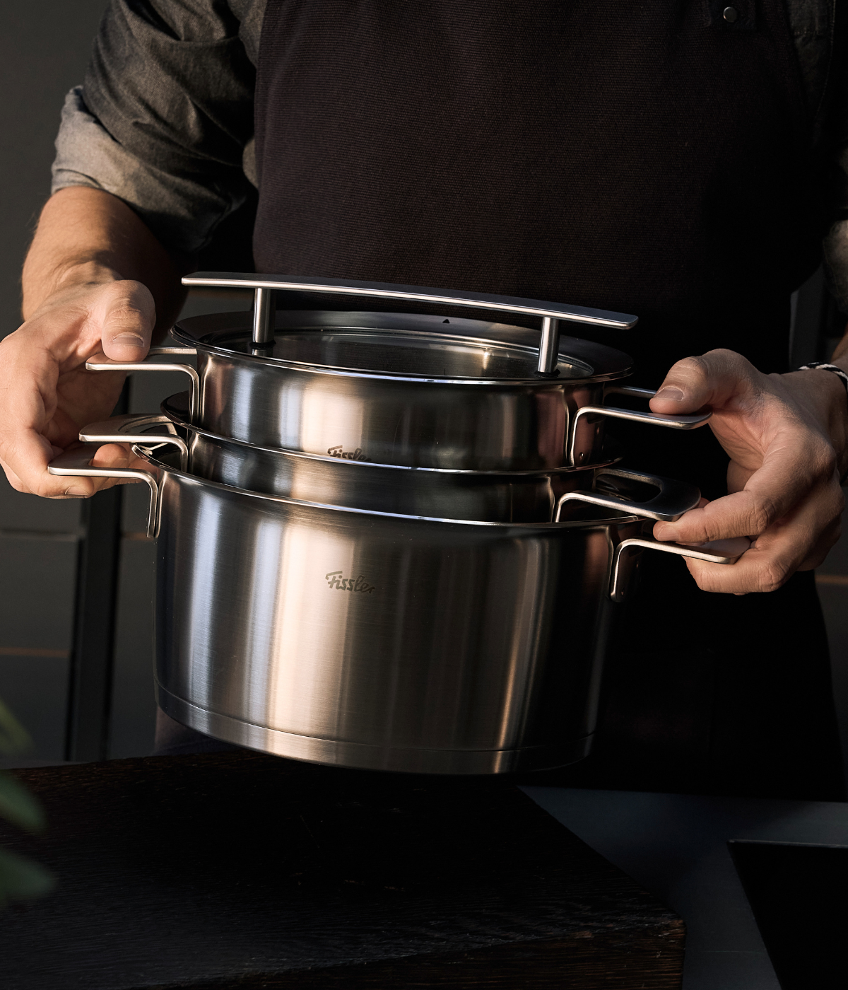 Eine Person in einer dunklen Schürze präsentiert zwei Fissler-Edelstahltöpfe mit ergonomischen Griffen, die elegant ineinander gestapelt sind und die Präzisionstechnik der Marke sowie das zeitlose deutsche Design in einer modernen Küchenumgebung widerspiegeln.