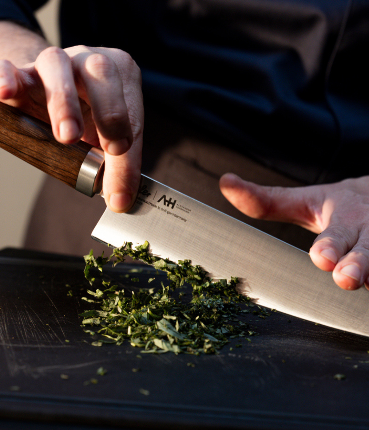 Eine Person schneidet frische Kräuter präzise auf einem eleganten schwarzen Schneidebrett mit einem hochwertigen Fissler-Küchenmesser mit einem elegant gearbeiteten Holzgriff – ein Spiegelbild des Bekenntnisses der Marke zu deutscher Ingenieurskunst, Langlebigkeit und zeitlosem Design.