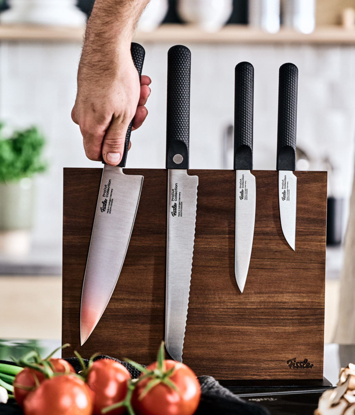 Eine Hand nimmt ein präzisionsgefertigtes Kochmesser aus einem Fissler-Holzblock und präsentiert damit vier vielseitige Fissler-Messer. Im Vordergrund unterstreichen frische Tomaten und duftende Kräuter das zeitlose Design und die deutsche Qualität, die den innovativen Küchenaccessoires von Fissler eigen sind.