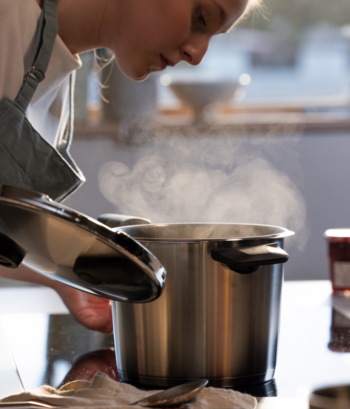In einer sonnendurchfluteten Küche hebt eine Person in einer Schürze vorsichtig den Deckel eines dampfenden Fissler-Edelstahltopfs an und präsentiert damit die für die Marke charakteristische Präzisionstechnik und das zeitlose deutsche Design. Sonnenlicht strömt durch das Fenster und beleuchtet das hochwertige Kochgeschirr, das das Herzstück der Heritage-Kollektion von Fissler bildet.