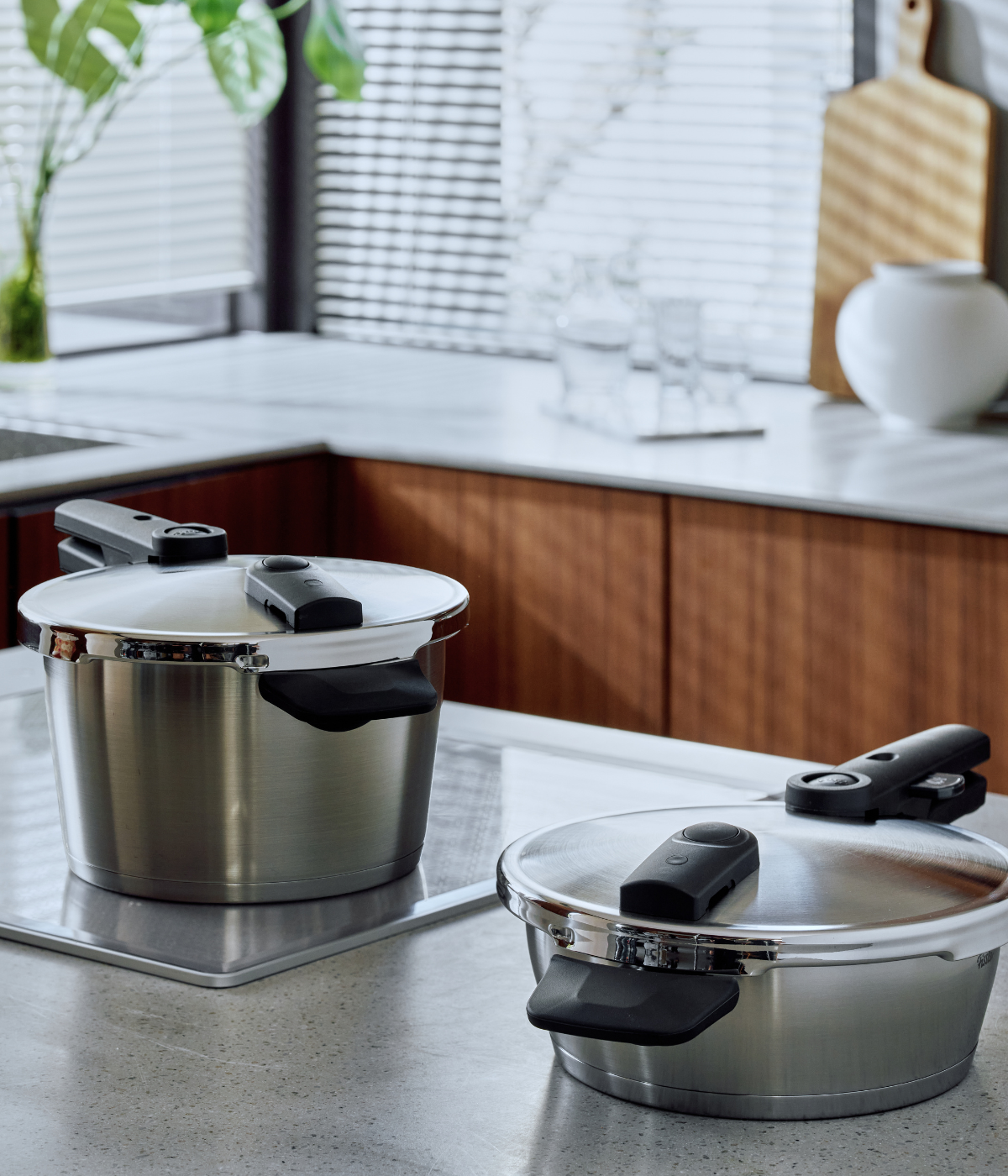 Zwei Fissler-Edelstahl-Schnellkochtöpfe mit den charakteristischen schwarzen Griffen stehen auf einer modernen Küchenarbeitsplatte. Sonnenlicht fällt durch die Jalousien, beleuchtet die Oberflächen aus weißem Marmor und unterstreicht die raffinierte Langlebigkeit sowie die präzise deutsche Ingenieurskunst dieser hochwertigen Kochgeschirrteile. Elegante Holzschränke bilden den Hintergrund und spiegeln Fisslers Tradition zeitlosen Designs und Innovationen seit 1845 wider.