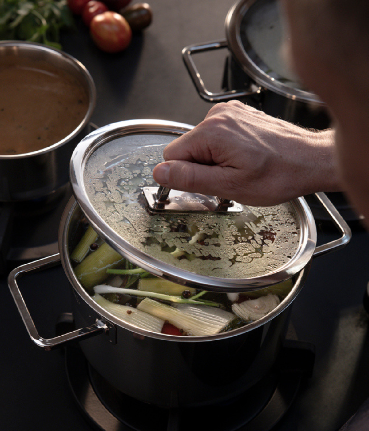 Eine Nahaufnahme aus der Vogelperspektive zeigt eine Hand, die den hochwertigen Glasdeckel von einem Fissler-Edelstahltopf abhebt, der fachmännisch für präzises Kochen entwickelt wurde. Der Topf steht auf einem modernen Herd und ist mit farbenfrohem Gemüse und einer würzigen Brühe gefüllt. Daneben steht ein weiterer glänzender Fissler-Topf, während frische Tomaten und Kräuter im sanft unscharfen Hintergrund farbliche Akzente setzen – ein Spiegelbild unserer Tradition innovativer deutscher Handwerkskunst
