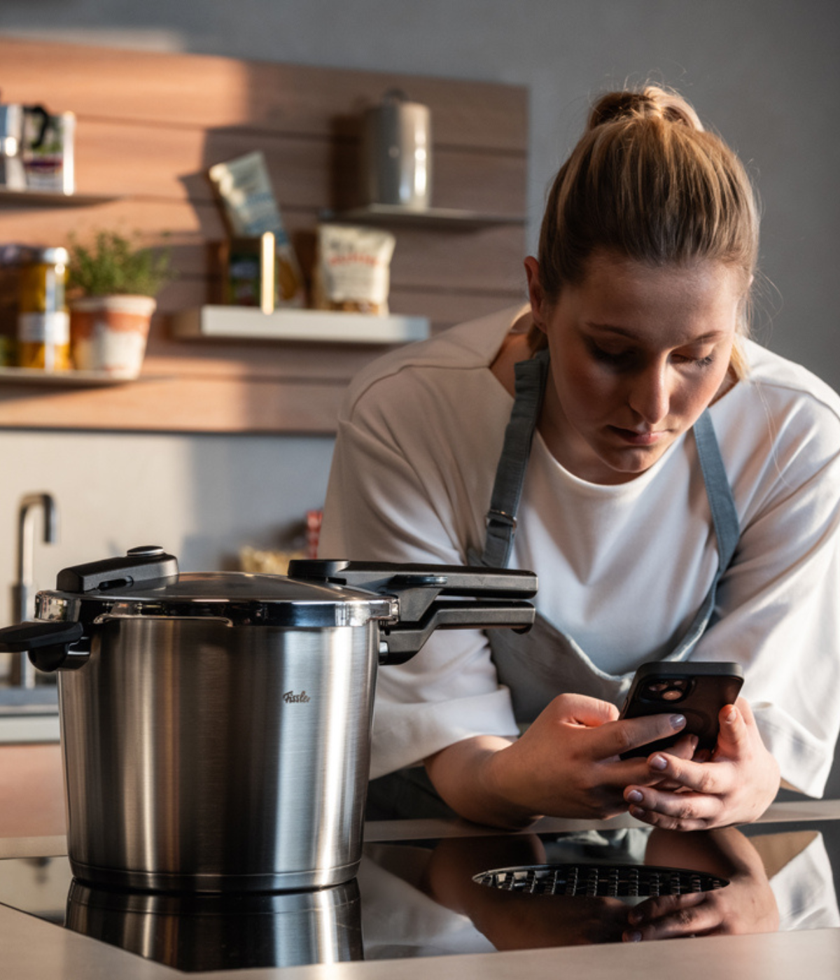 Eine Frau in einer Schürze lehnt an der Küchentheke und schaut auf ihr Handy, das neben einem hochwertigen Fissler-Edelstahl-Schnellkochtopf auf dem Herd liegt. Regale mit ordentlich aufgereihten Gläsern und Zutaten unterstreichen die gut ausgestattete Küche und spiegeln die Tradition von Fissler wider: deutsche Ingenieurskunst, zeitloses Design und langlebiges Kochgeschirr.