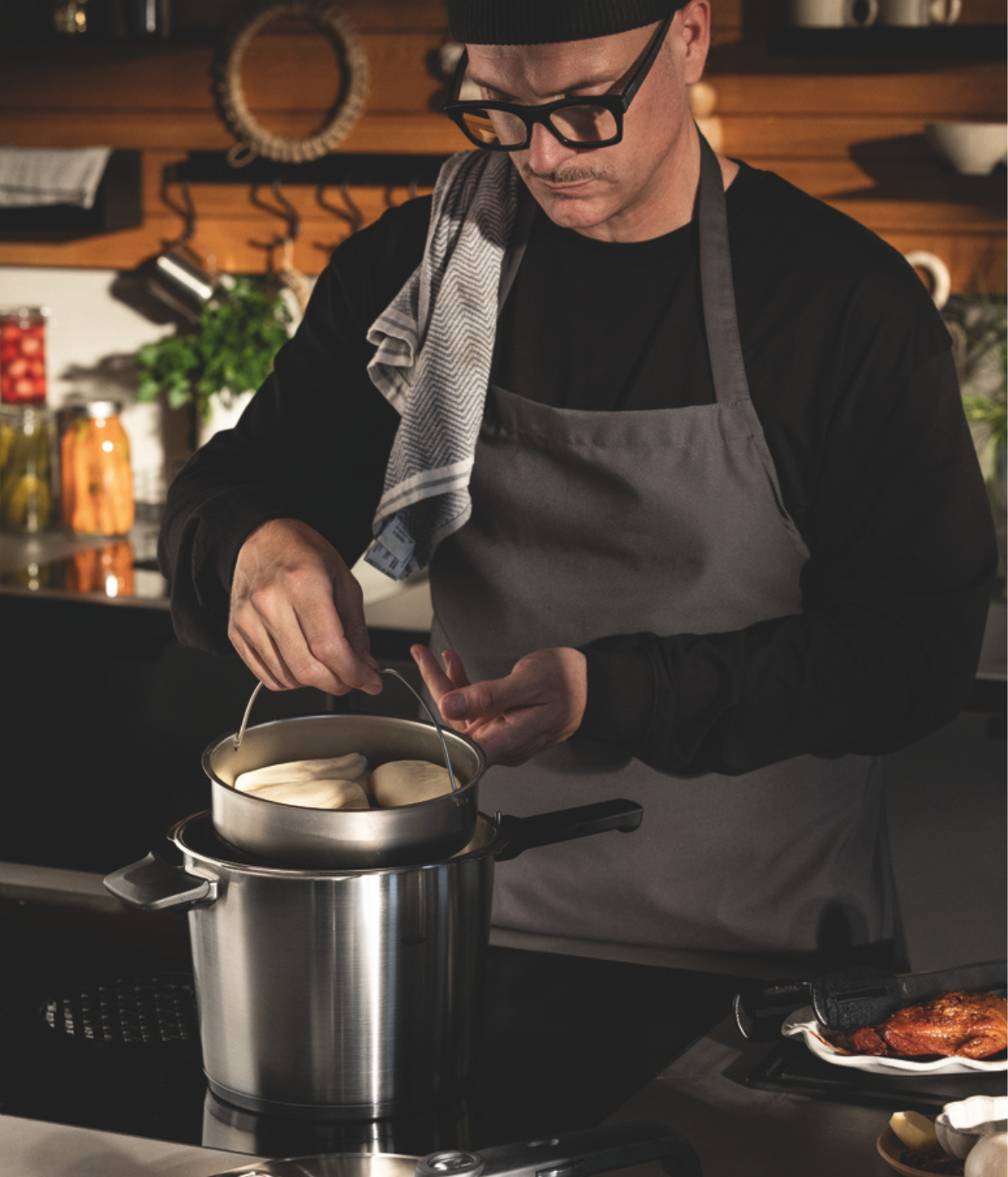 In einer gut ausgestatteten Küche dämpft ein Mann, der eine Brille, einen Hut und eine Schürze trägt, gekonnt ein Tablett mit Brötchen über einem hochwertigen Fissler-Edelstahltopf. Mit Gläsern gefüllte Regale bilden eine einladende Kulisse und unterstreichen die präzise Technik und das zeitlose Design der Fissler-Kochgeschirre, die im Mittelpunkt des Kochvorgangs stehen.