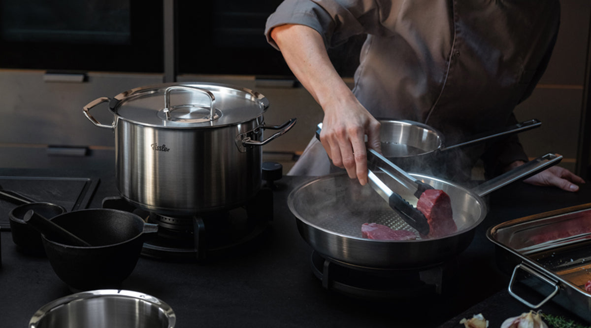 Ein Koch in weißem Kittel brät gekonnt ein Stück Fleisch mit einer Zange in einer hochwertigen Fissler-Edelstahlpfanne auf dem Herd an, während ein passender Fissler-Topf und hochwertige Küchenutensilien, die für deutsche Präzision und Innovation bekannt sind, griffbereit bereitstehen.